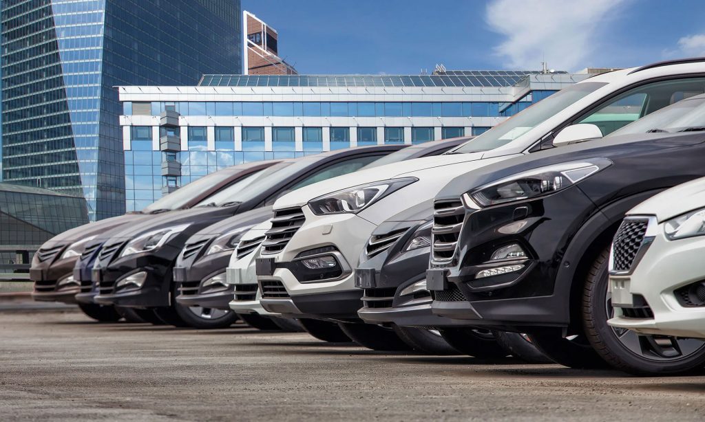 A row of parked cars in a parking lot with modern buildings in the background.
